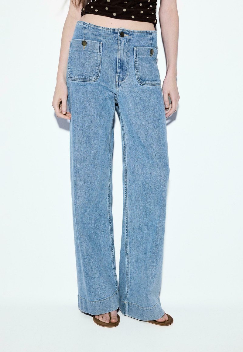 Femme portant un jean denim bleu clair à jambes larges avec de grandes poches plaquées devant et des sandales marron, debout sur fond blanc.