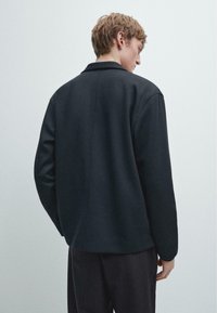 Giacca casual nera con taglio dritto, dotata di colletto, maniche lunghe e texture liscia. La vista posteriore mostra dettagli minimi nelle cuciture.