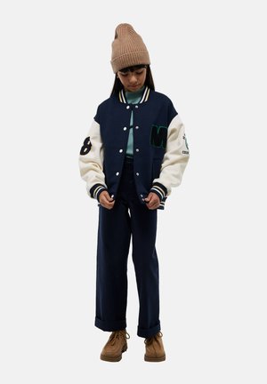 Veste varsity bleu marine et crème avec des patches de lettres et de chiffres verts. Portée sur un pull léger, associée à un pantalon bleu marine et des chaussures marron.
