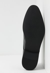 Semelle de chaussure noire avec un fond en caoutchouc texturé présentant des motifs en zigzag. Talon rigide avec une surface supérieure lisse et brillante.