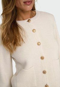 Cardigan a maglia crema con maniche lunghe, caratterizzato da un collo rotondo, due tasche anteriori e quattro bottoni dorati per la chiusura.