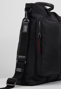 Sac en tissu noir avec bandoulière réglable, poche zippée à l'avant, détails de surpiqûre rouge sur la fermeture éclair et poignée de transport supérieure, sur fond gris.