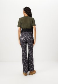 Haut à manches courtes vert olive tricoté, associé à un pantalon noir évasé avec un motif floral blanc. Des chaussures compensées marron complètent la tenue.