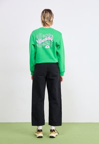 Grön sweatshirt med rosa grafisk text på ryggen, tillsammans med svarta vida byxor och beiga sneakers. Texturerad bomullstyg.