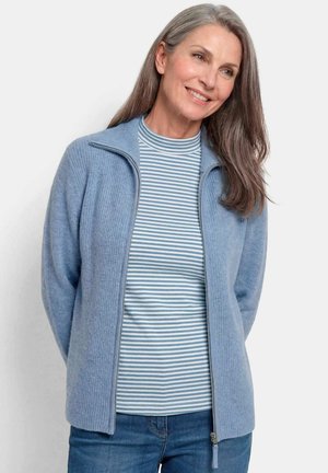 Donna matura sorridente con capelli lunghi e grigi che indossa un maglione azzurro con zip sopra una camicia a righe bianche e blu e jeans blu.