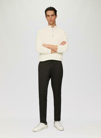 Witte ritsvest, zwarte broek met aansnoerkoord en witte sneakers met zwarte accenten. Model staat met gekruiste armen tegen een lichte achtergrond.