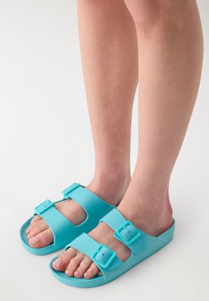 Piedi che indossano sandali turchesi con punta aperta e due fibbie regolabili su sfondo bianco.
