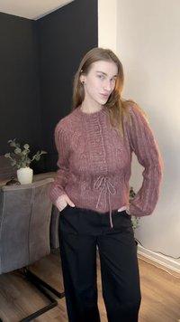 Πλεκτό πουλόβερ με πλέξη καλώδιο σε σκουρό ροζ απόχρωση, με στρογγυλή λαιμόκοψη, λεπτομέρεια με κουμπιά και σκοινί στη μέση, συνδυασμένο με μαύρο παντελόνι φαρδιάς γραμμής.