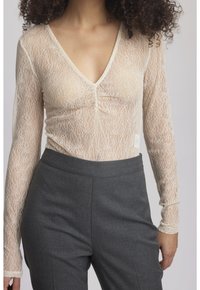 Vrouw in een doorschijnende crèmekleurige kanten langemouwen top met een geplooide V-hals, gecombineerd met hoog-gewijde donkergrijze broek.