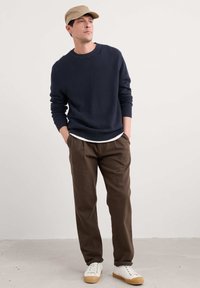 Marinblå strukturerad tröja, vit t-shirt, bruna veckade byxor och vita sneakers med bruna sulor. Bärs med en beige keps.