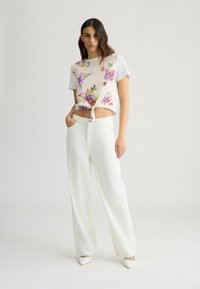 Hvid blomstret crop top med bindebånd, parret med løse hvide bukser. Modellen står med hænderne i lommerne på en ensfarvet baggrund.