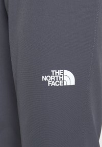 Szary materiał o gładkiej teksturze, z białym logo "The North Face" w dolnym rogu oraz prostymi detalami przeszycia.