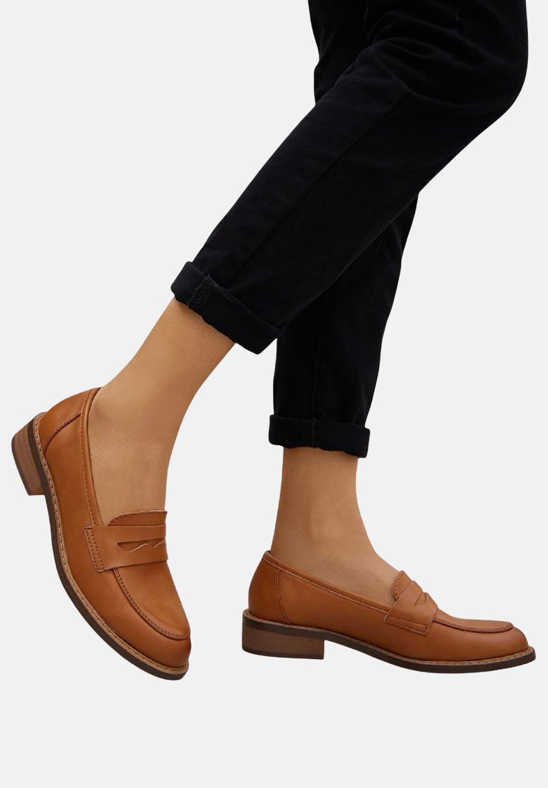 Mocassins en cuir marron avec un petit talon carré, présentant un design cousu et un détail de sangle à l'avant. Associés à un pantalon noir retroussé.