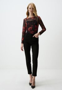 Un pantalon noir taille haute associé à un haut transparent à manches longues avec des motifs floraux rouges sur un fond noir. Des escarpins noirs à talons pointus complètent le look.