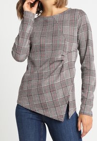 Haut gris à manches longues avec motif à carreaux rouges et noirs, présentant un ourlet asymétrique et un tissu texturé. Associé à un jean bleu.