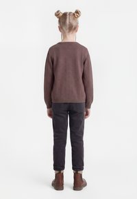 Barn med blondt hår sat op i to knolde iført en brun sweater, mørke bukser og brune støvler, stående med ryggen mod på en hvid baggrund.