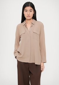 Bluza bej cu mâneci lungi, cu închidere cu nasturi în față, două buzunare la piept și croială lejeră. Textură netedă și luciu subtil, asortată cu pantaloni maro.