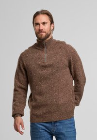 Brauner Strickpullover mit hohem Kragen, halbem Reißverschluss und strukturierter Oberfläche. Getragen über blauen Jeans mit einem feinen gesprenkelten Muster.