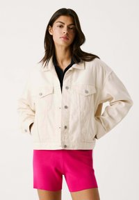 Chaqueta de mezclilla crema con un corte relajado, dos bolsillos frontales, botones plateados y una forma ligeramente oversized. Llevada sobre una camiseta azul marino y pantalones cortos rosas.
