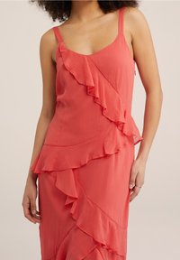 Coral dress with a pleated fabric, featuring ruffles along one side and thin straps. The design is form-fitting and elegant.