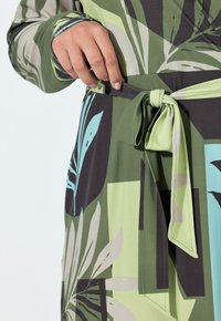 Zielono-czarna sukienka z dużymi wzorami liści, związaną paskiem w talii i długimi rękawami z kontrastowymi mankietami. Gładka faktura materiału.