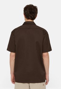 Muž s tmavě hnědým polokošilí krátkým rukávem a límečkem, viděný zezadu, v kombinaci se světle béžovými šortkami.