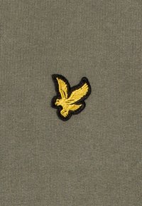 Żółty haftowany ptak w locie z czarnym konturem na oliwkowej, teksturowanej tkaninie.