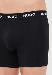 Boxeri negri dintr-un material neted, cu un tiv elastic branduit pe care este imprimat "HUGO" în alb.