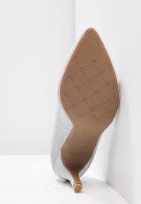 Šedá semišová lodička na vysokém podpatku s špičatou špičkou, která má béžovou gumovou podrážku s vyraženým vzorem srdce a mírným podpatkem.