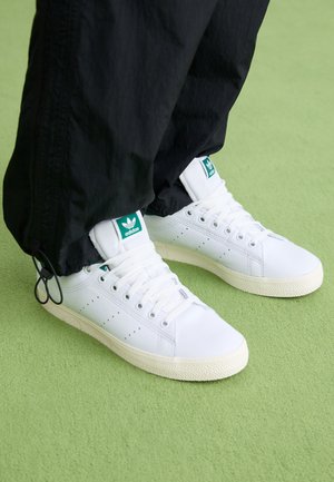 Hvide Adidas sneakers med grønt logo, båret med sorte bukser på lysegrøn gulv.
