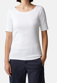 Bílý krátký rukáv top s širokým výstřihem a ohrnutými manžetami, vyrobený z měkké tkaniny; spárován s tmavě námořnickými kalhotami, ruce v kapsách.