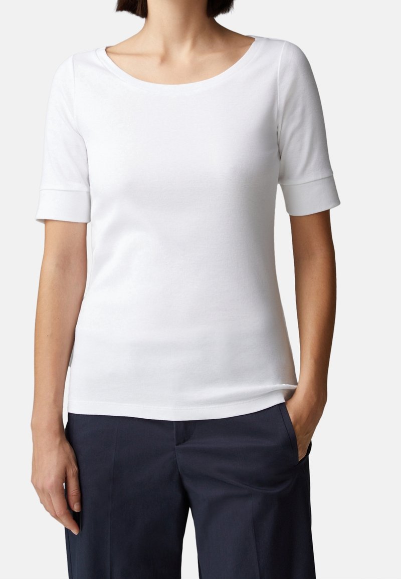 Bílý krátký rukáv top s širokým výstřihem a ohrnutými manžetami, vyrobený z měkké tkaniny; spárován s tmavě námořnickými kalhotami, ruce v kapsách.