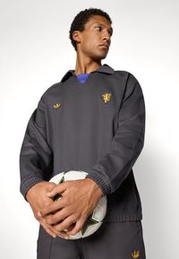 Juodas sportinis megztinis su apykakle ir mažu auksiniu logotipu. Modelis laiko baltą futbolo kamuolį su juodais ir geltonais akcentais.