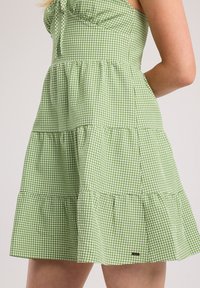 Vihreä ruudullinen mekko kerroksellisella designilla, olkaimilla ja pienellä logokoristeella. Puuvillakangasta, jossa on teksturoitu pinta.