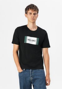 Svart bomull T-skjorte med et grønt og hvitt rektangulært logo med "JACK & JONES." Modellen har på seg denimjeans med hendene i lommene.