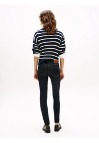Mujer de pie de espaldas con un suéter a rayas azul marino, camisa blanca con las mangas arremangadas, jeans oscuros ajustados y zapatos negros sobre un fondo blanco.