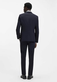 Abito blu navy con vestibilità su misura, presenta una singola spaccatura posteriore e pantaloni a pieghe piatte, completato da scarpe nere e una camicia bianca.