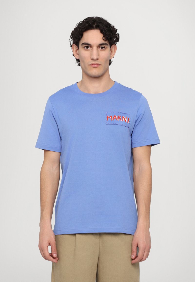 T-shirt en coton bleu clair à manches courtes, col rond, avec un logo rouge brodé sur un patch sur la poitrine gauche.