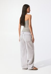 Jasnoszare dżinsy typu wide-leg z charakterystycznymi tylnymi kieszeniami, w połączeniu z dopasowaną oliwkową bluzką na ramiączkach. Modelka stoi w profilu, w sandałach.