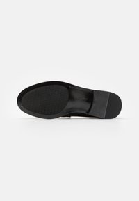 Semelle de chaussure noire avec une zone avant texturée pour l’adhérence, une section médiane lisse avec logo, et un talon solide, présentée sur un fond blanc uni.