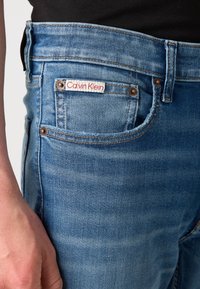 Gaiši zili džinsi ar priekšējo kabatu, zeltītām šuvēm un redzamu Calvin Klein uzrakstu. Gluda tekstūra ar klasisku piegriezumu.