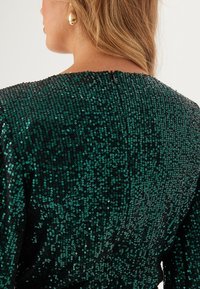 Abito verde con paillettes su base nera, presenta un collo alto e chiusura con zip. Texture liscia con paillettes riflettenti che creano un effetto scintillante.