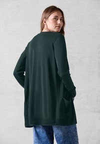 Mörkgrön långärmad cardigan i mjukt tyg. Har en avslappnad passform, sidofickor och ribbade ärmslut och fåll.