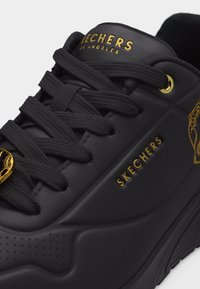 Μαύρο αθλητικό παπούτσι Skechers με παχιά σόλα, χρυσό λογότυπο στο πλάι και στη γλώσσα, μαύκα κορδόνια και λεπτομέρειες σε χρυσή τρύπα κορδονιού.