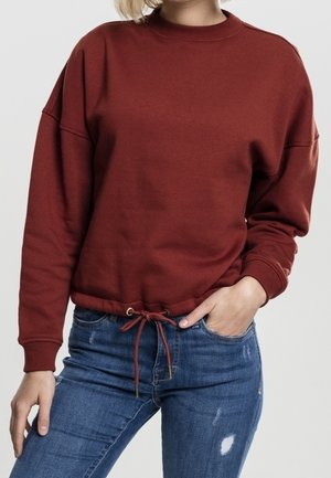 Personne portant un sweat-shirt couleur rouille avec un cordon au bas et un jean bleu déchiré, une main dans la poche, sur un fond uni.