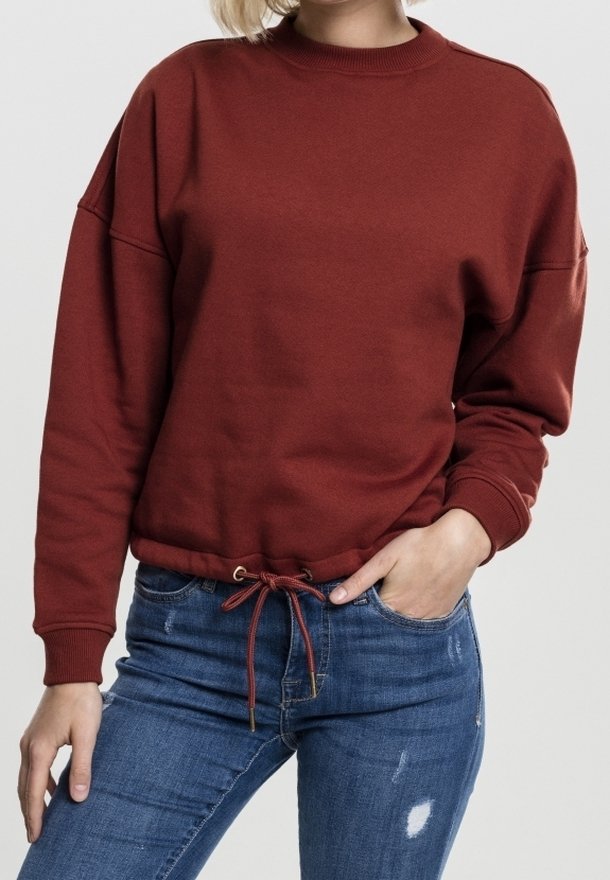 Personne portant un sweat-shirt couleur rouille avec un cordon au bas et un jean bleu déchiré, une main dans la poche, sur un fond uni.