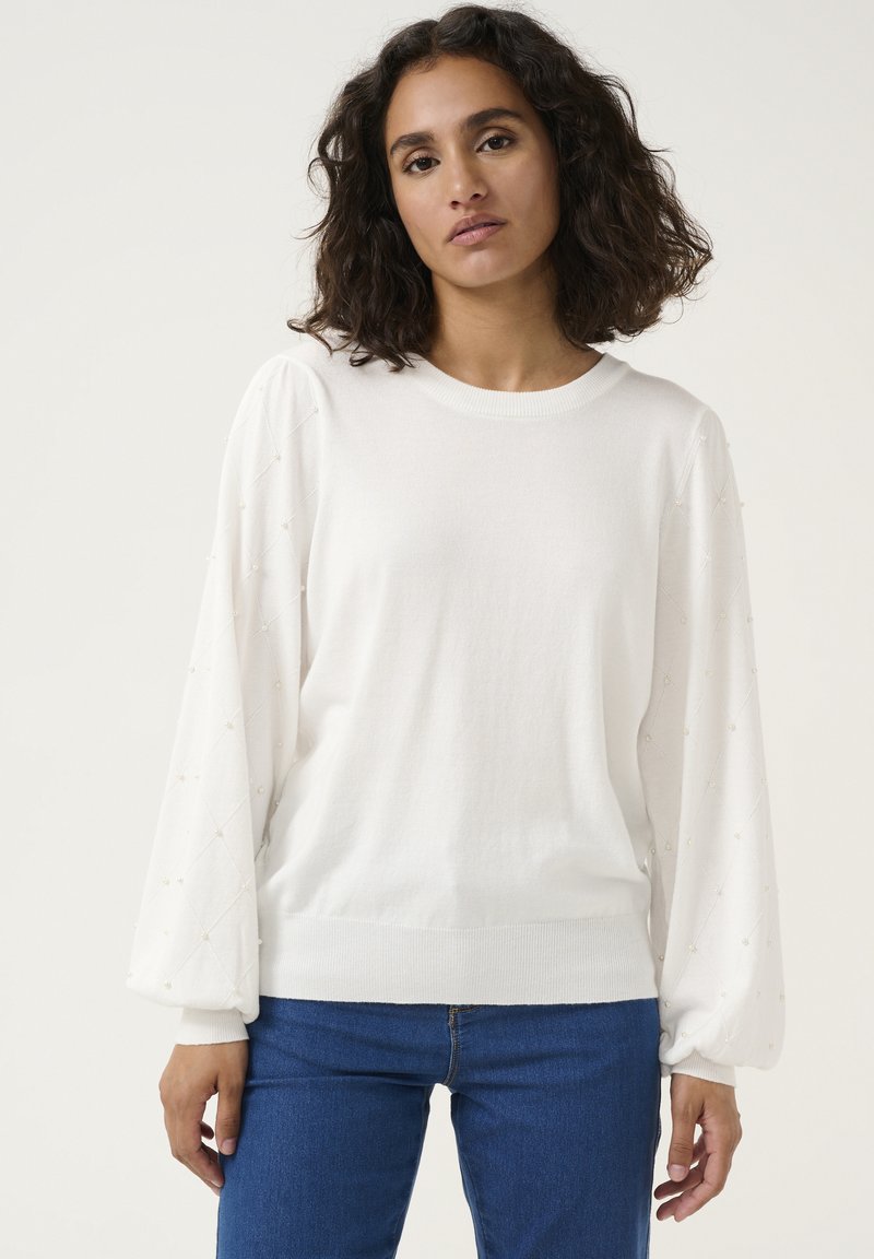 Sweater blanc en tricot avec des manches bouffantes, présentant des accents texturés et de subtils détails en perles sur les manches. Associé à un jean bleu.