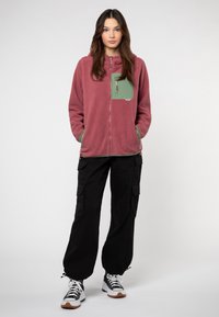 Fleece jack met rits met een roze lijf en een groene borstzak, gecombineerd met zwarte cargo-broeken en zwart-witte sneakers.