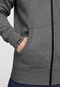 Grå zip-up hoodie med ribbede manchetter og kant. Har en frontlomme og et lille logo-tag på manchetten. Glat tekstur, afslappet design.