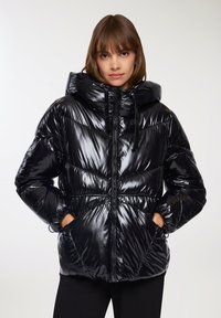 Giacca a vento nera con superficie lucida e trapuntata, cappuccio oversize, zip frontale e due tasche laterali; presenta un design con vita arricciata.